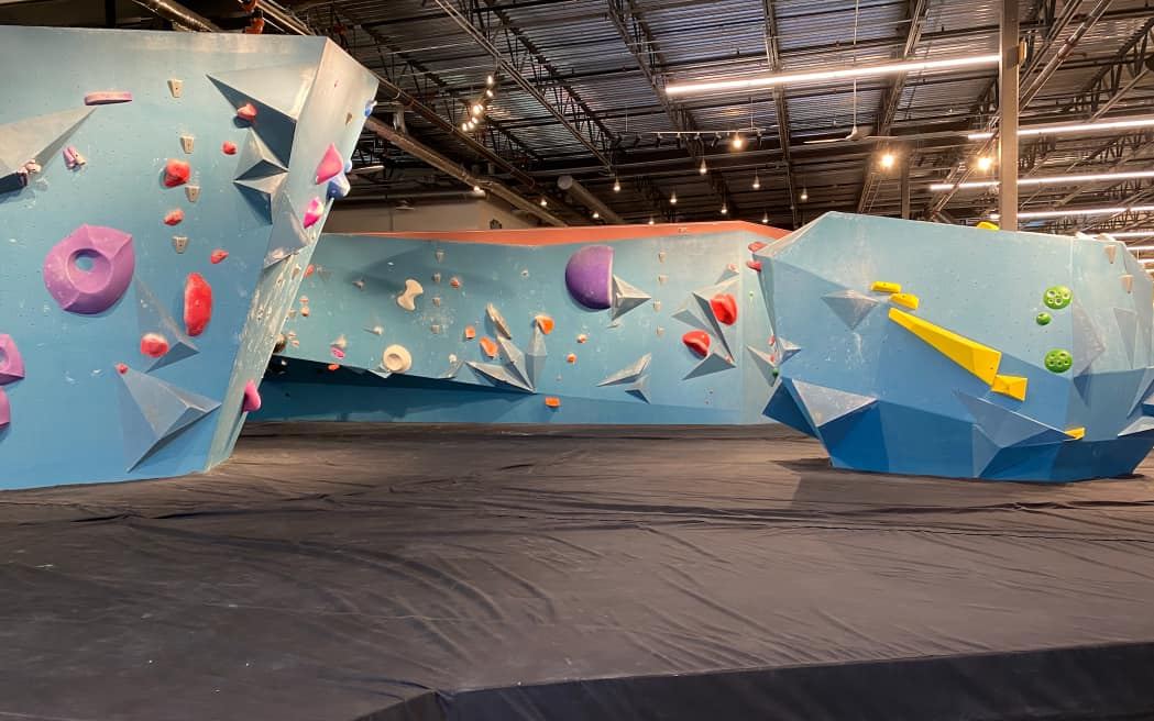 Blue freestanding bouldering walls with angled volumes and holds, over padded flooring in a warehouse gym