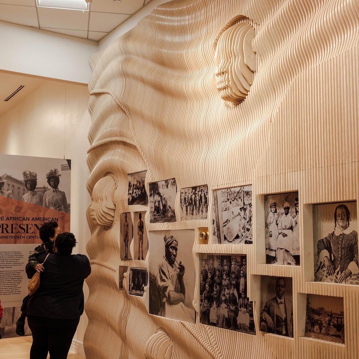 Full view of the wall with a large sliced head in relief above a grid of archival photographs, beside the exhibit title banner