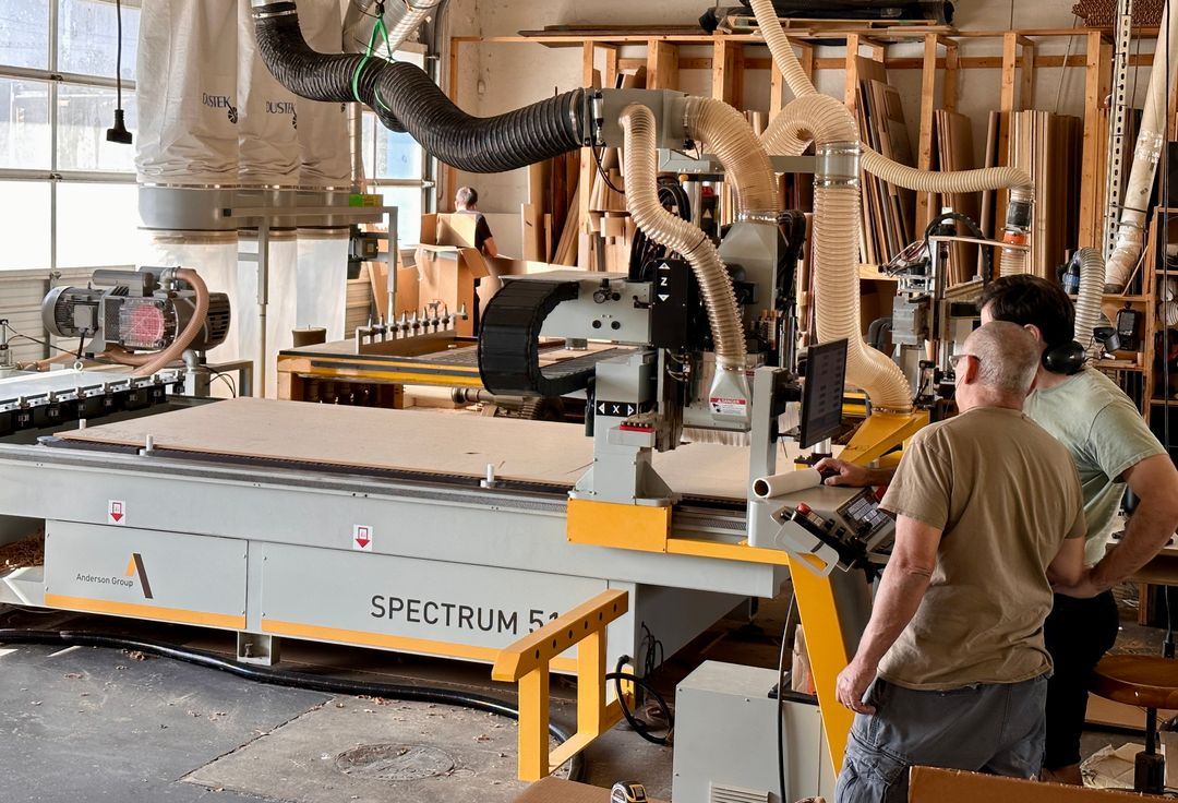 Two people at the controls of an Anderson Spectrum CNC router with a sheet on the bed and dust collection hoses overhead