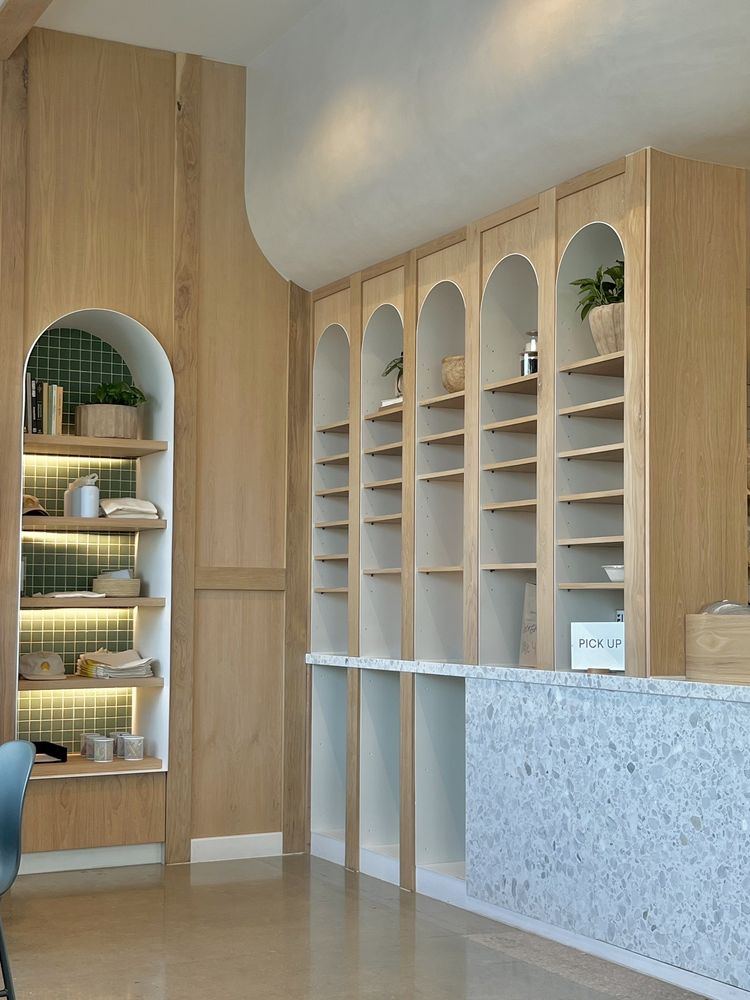 Arched open shelving in white oak above a terrazzo pickup counter, next to a tiled niche with lit shelves