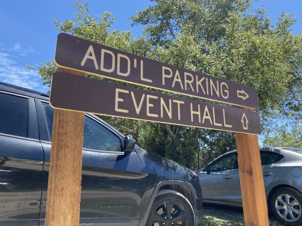 Carved sign reading Add'l parking and Event hall with arrows, on wood posts beside parked cars