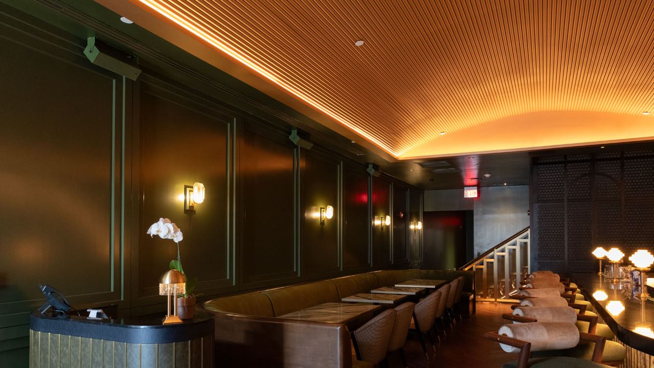Dark green paneled walls with brass sconces above a long banquette, under a curved slatted ceiling