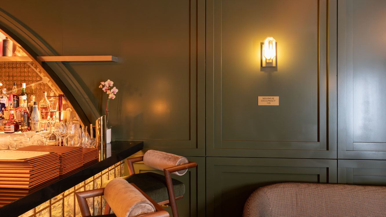 Green painted wall panels with applied moulding next to the arched back bar, with a sconce and two bar stools