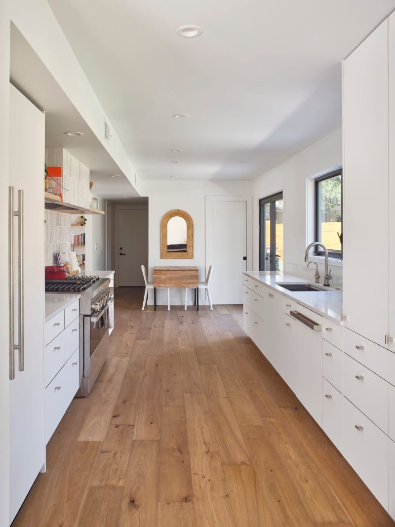 View down the kitchen between two runs of flat white cabinets with long pulls, toward an arched mirror