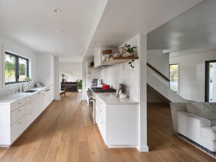 Kitchen with white cabinets along the window wall and a range wall with a floating wood shelf, open to the living room and stairs