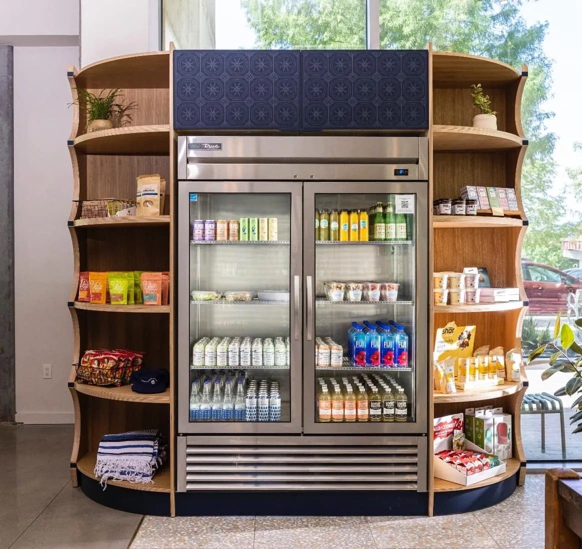 Glass-door reach-in cooler framed by curved wood shelving stocked with snacks, with a navy patterned panel above