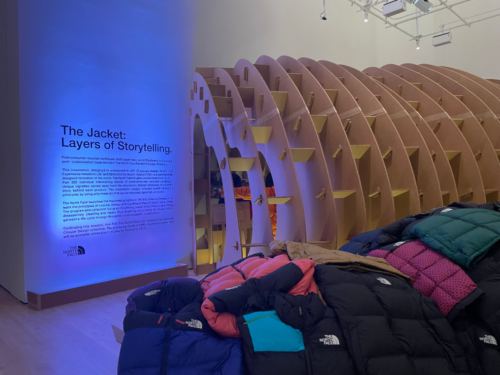 Cardboard ribs with small shelves rising behind a pile of The North Face jackets, next to the lit wall text