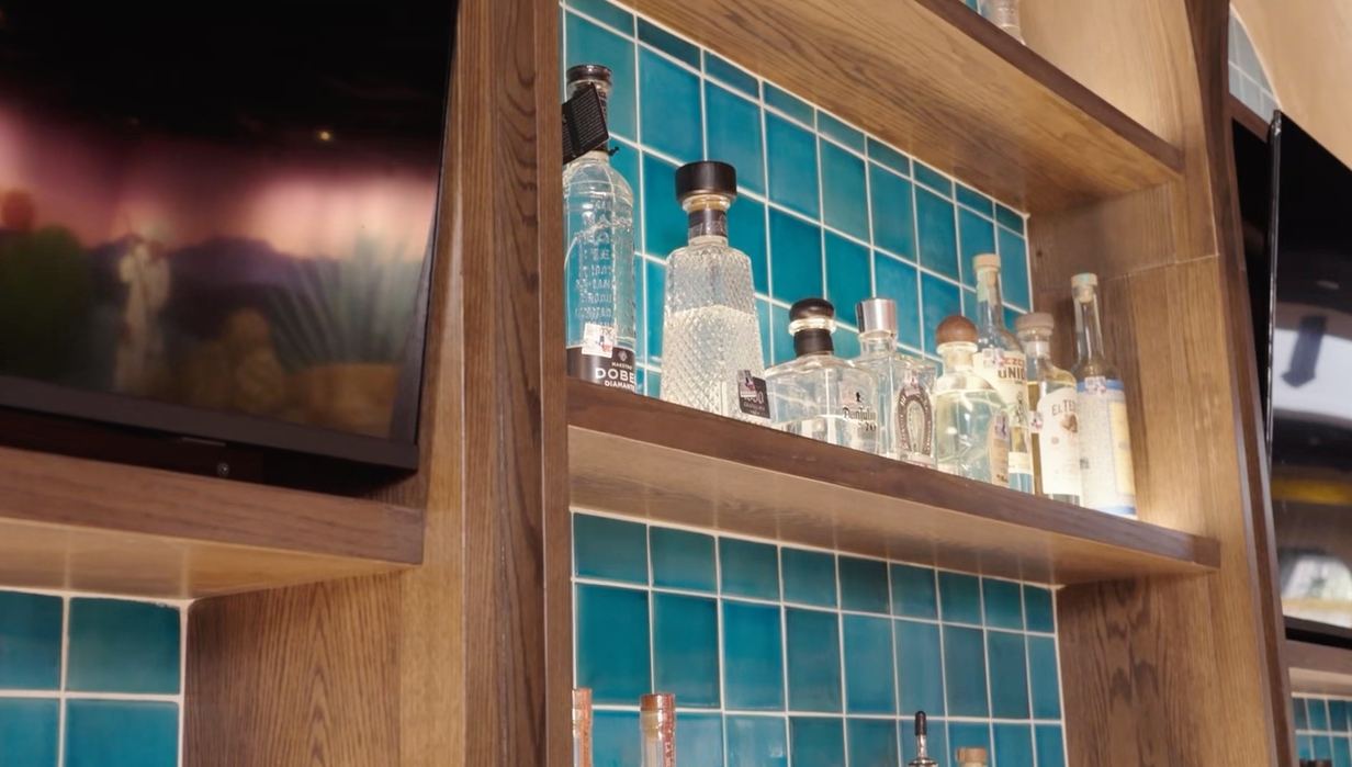 Wood bar shelving holding tequila bottles against turquoise tile, next to a mounted TV